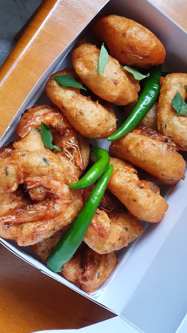 A box filled with golden-brown fried snacks, including shrimp fritters, garnished with green chilies and curry leaves.