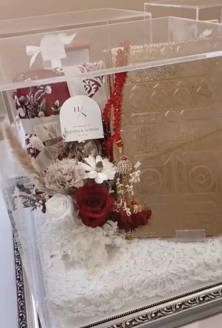 A beautifully arranged Islamic gift set featuring a golden Quran, prayer mat, tasbih, and floral decor in a clear display box.
