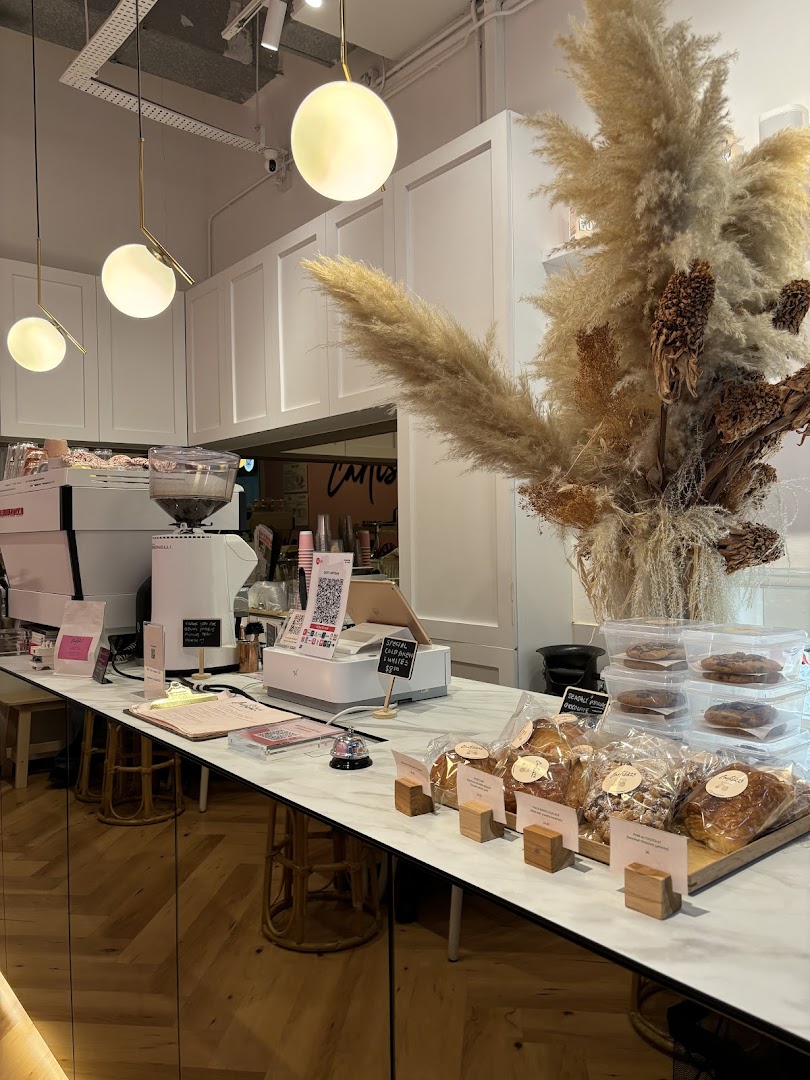 A well-lit coffee shop counter featuring coffee machines, pastries, and decorative pampas grass.