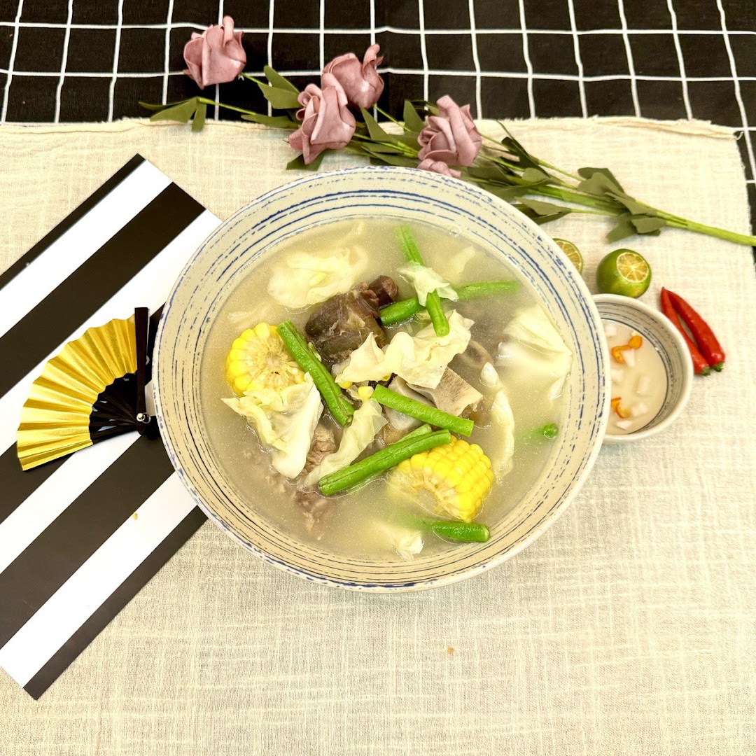 A hearty bowl of soup with meat, corn, green beans, and cabbage, served with lime and chili.