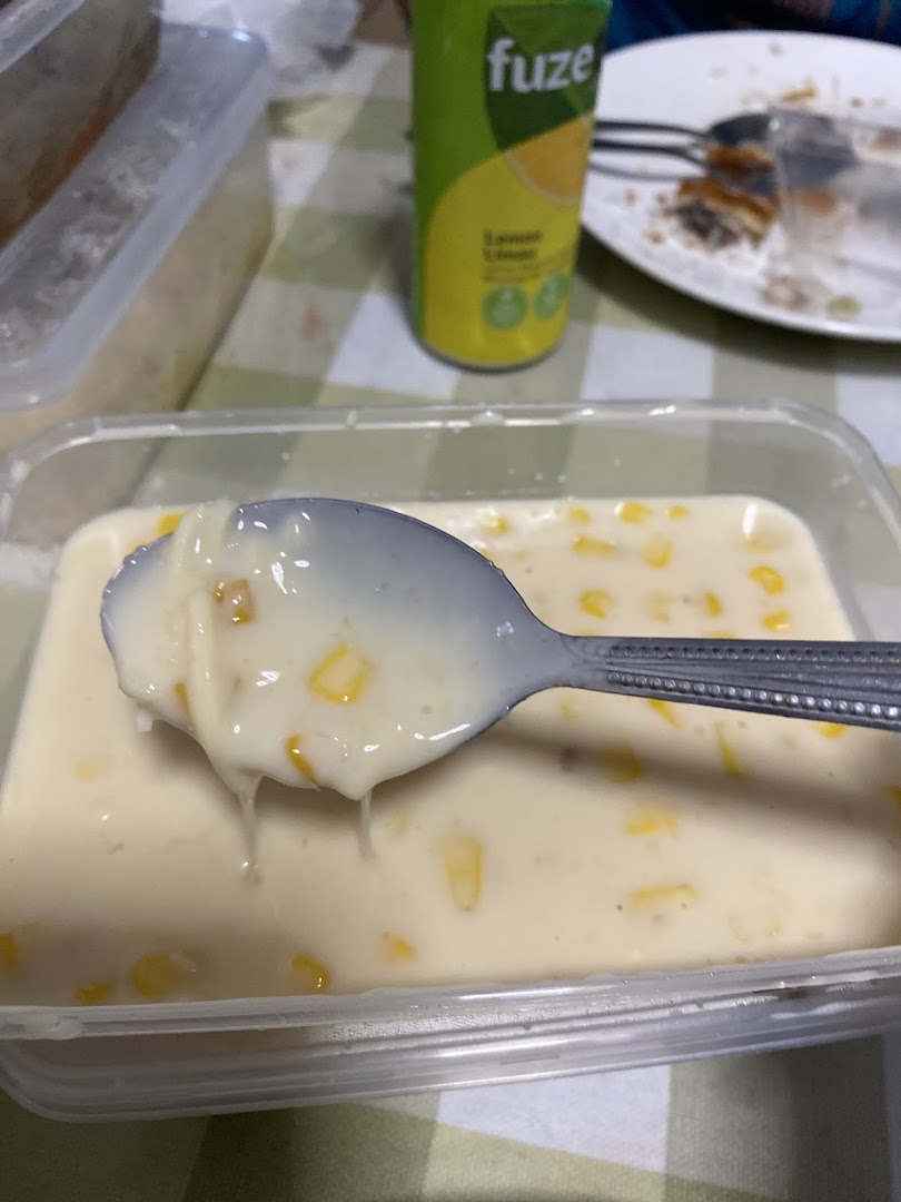 A spoon lifts creamy corn dessert from a clear container, with a Fuze drink can and plate in the background.