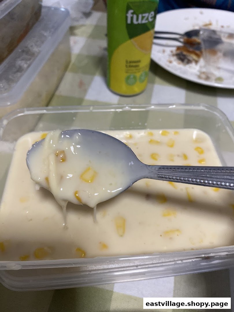 A spoon lifts creamy corn dessert from a clear container, with a Fuze drink can and plate in the background.