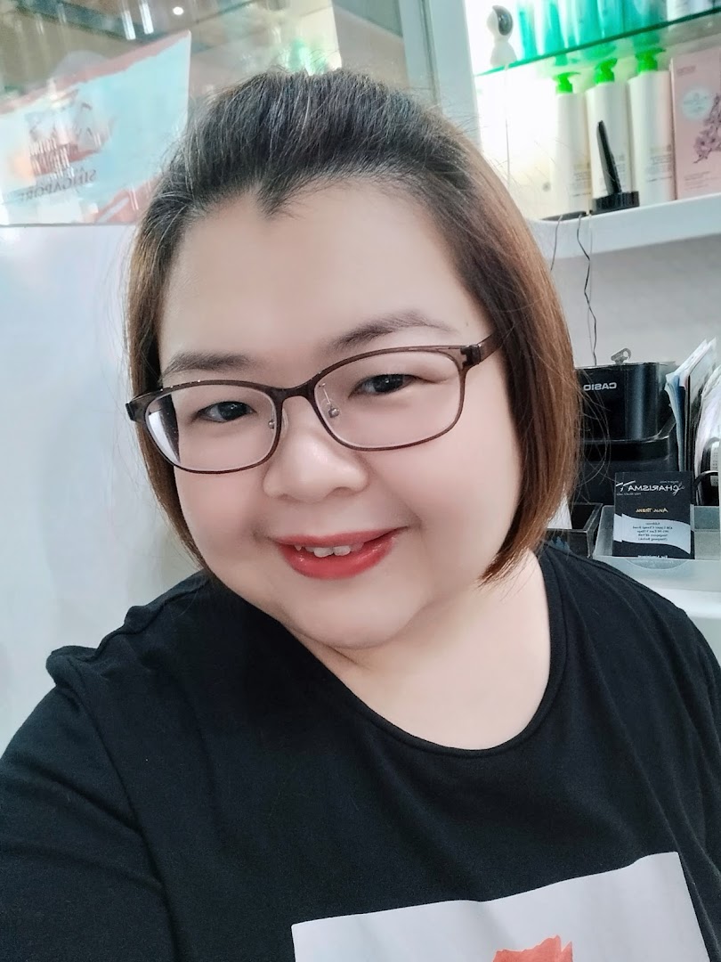 A smiling person wearing glasses, likely staff, inside Charisma Hair Studio with hair products in the background.