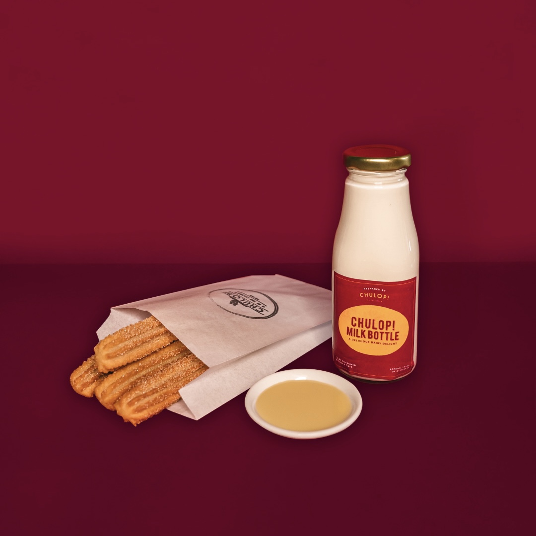 A paper bag of churros with a dipping sauce and a Chulop! Milk Bottle on a red background.