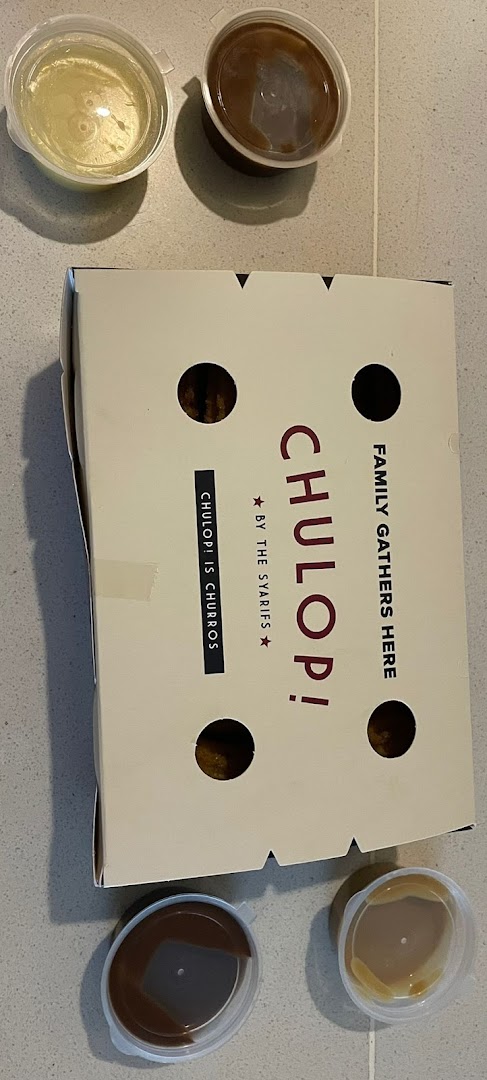 A box of Chulop! churros with four dipping sauces on a tiled surface.
