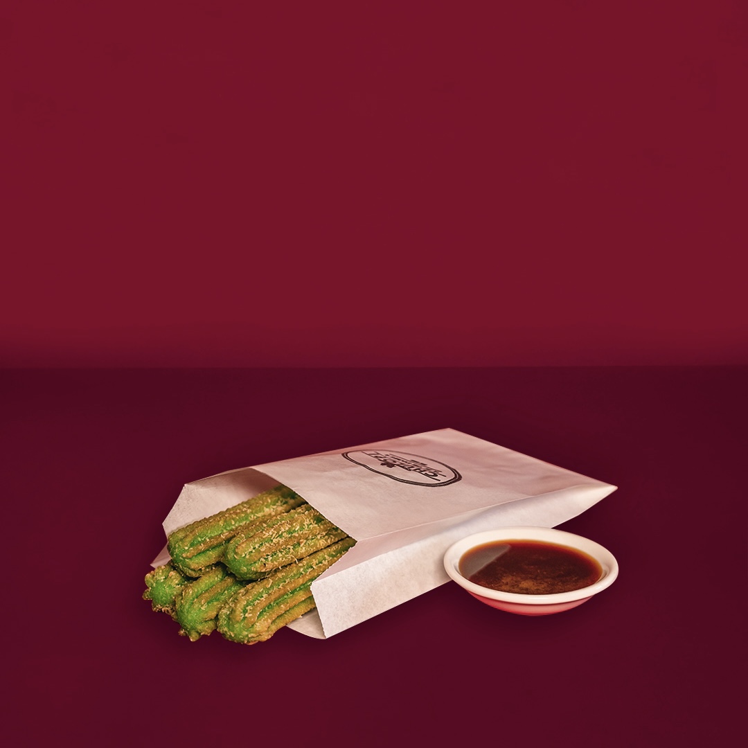 Vibrant green churros in a branded paper bag, served with a rich dipping sauce.