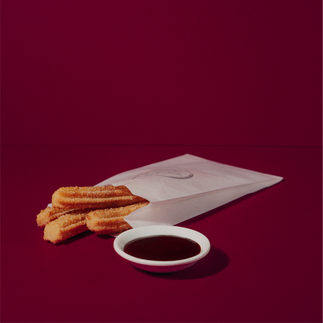 Golden churros in a paper bag with a side of rich chocolate dipping sauce.