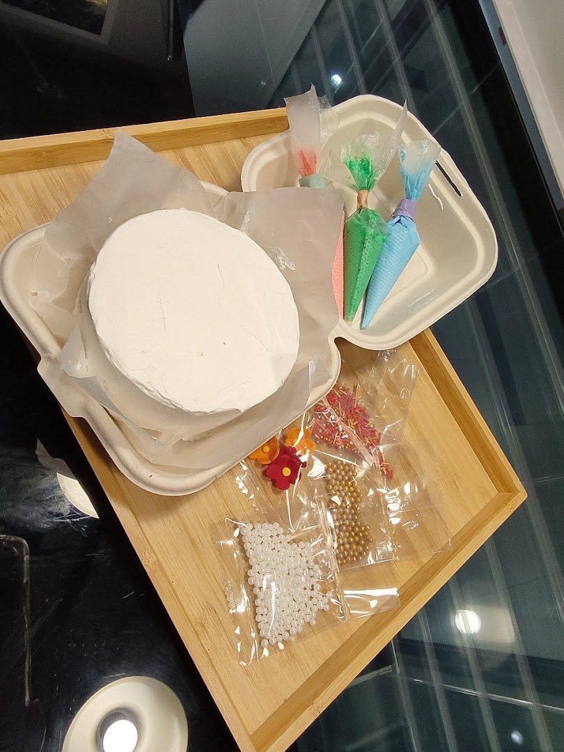 A DIY cake decorating kit featuring a plain cake, frosting bags, and colorful sprinkles on a wooden tray.