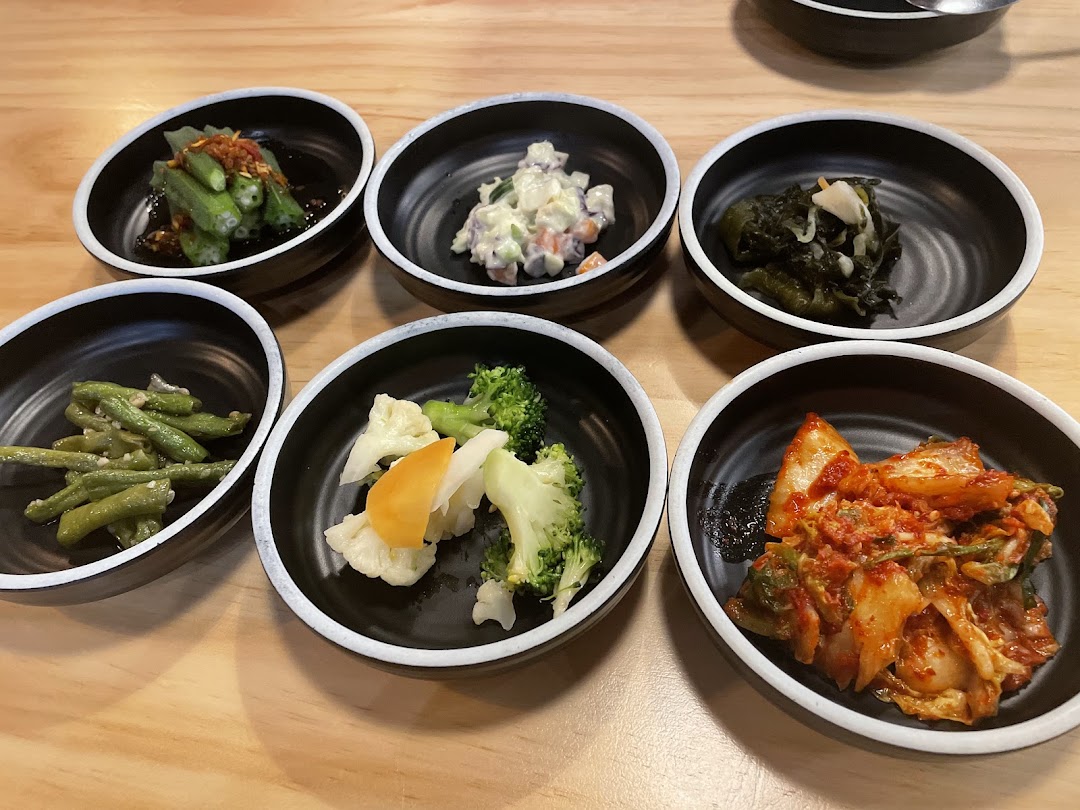 A variety of colorful Korean banchan (side dishes) served in small black bowls on a wooden table.
