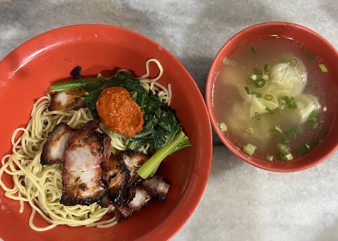 A vibrant bowl of char siu noodles with greens and chili paste, accompanied by a clear wanton soup.