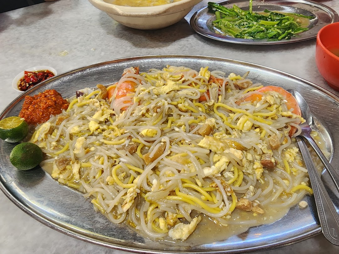 A close-up of a plate of stir-fried noodles with prawns, eggs, and lime, served with chili paste.