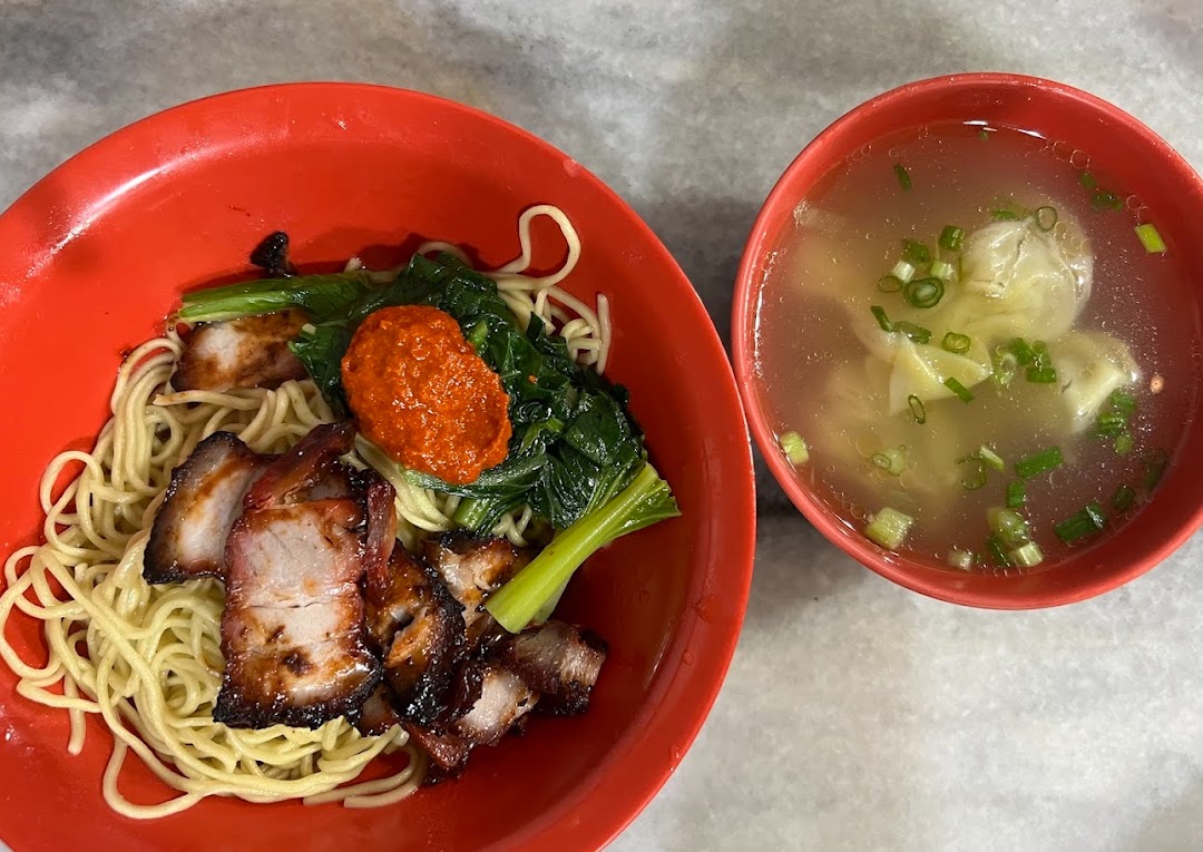 A vibrant red bowl of char siu noodles with greens and chili, accompanied by a clear wonton soup.
