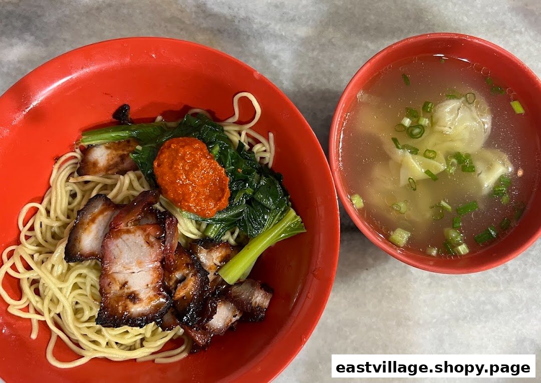 A vibrant red bowl of char siu noodles with greens and chili, accompanied by a clear wonton soup.