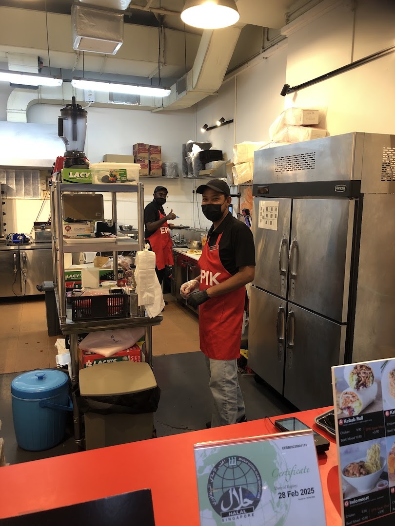 Two staff members in red aprons working in a clean kitchen, with a halal certificate visible on the counter.