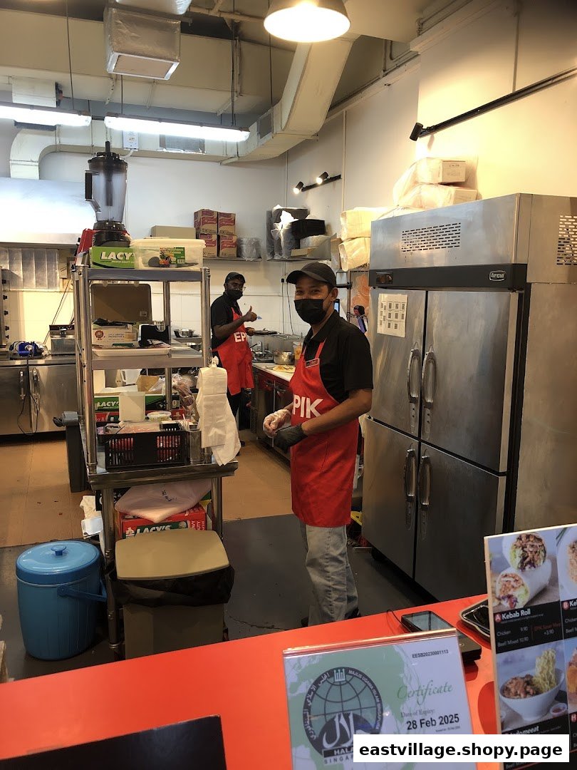 Two staff members in red aprons working in a clean kitchen, with a halal certificate visible on the counter.