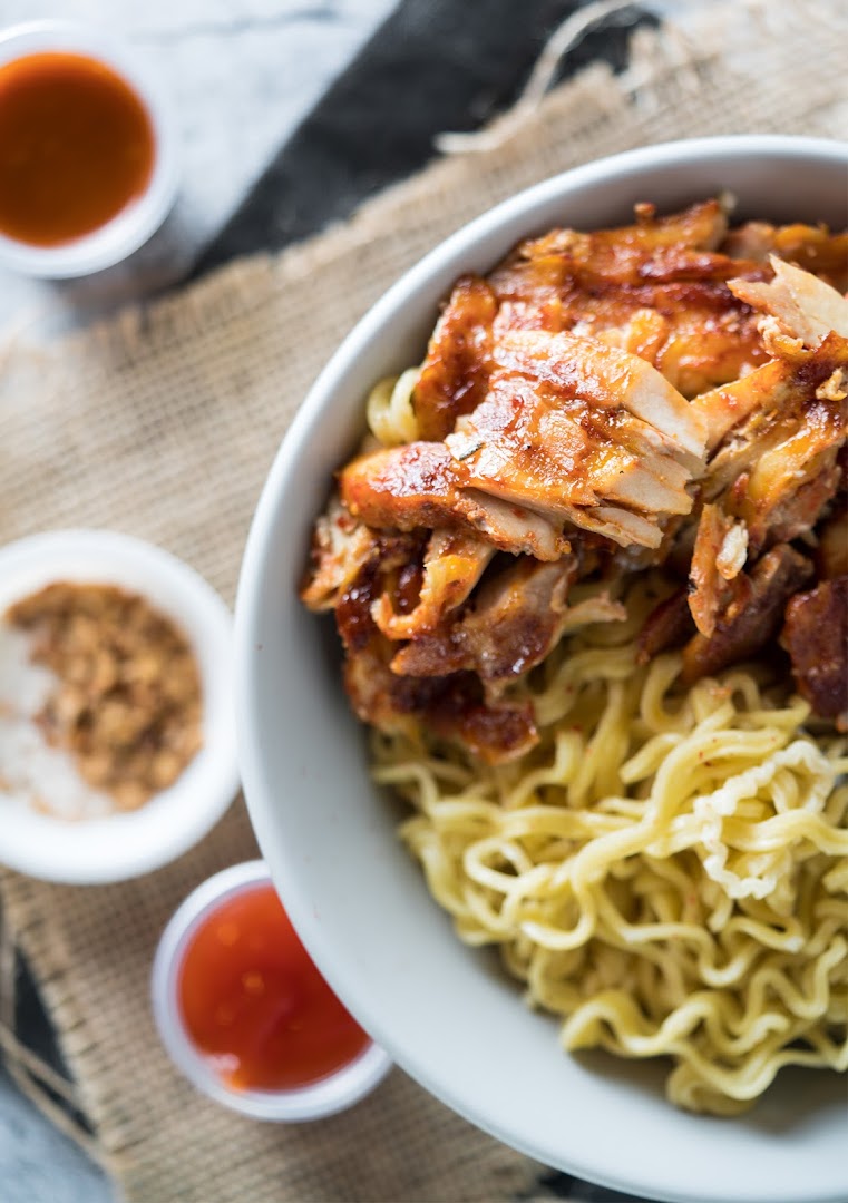 A close-up of a bowl of noodles topped with grilled chicken and side sauces.