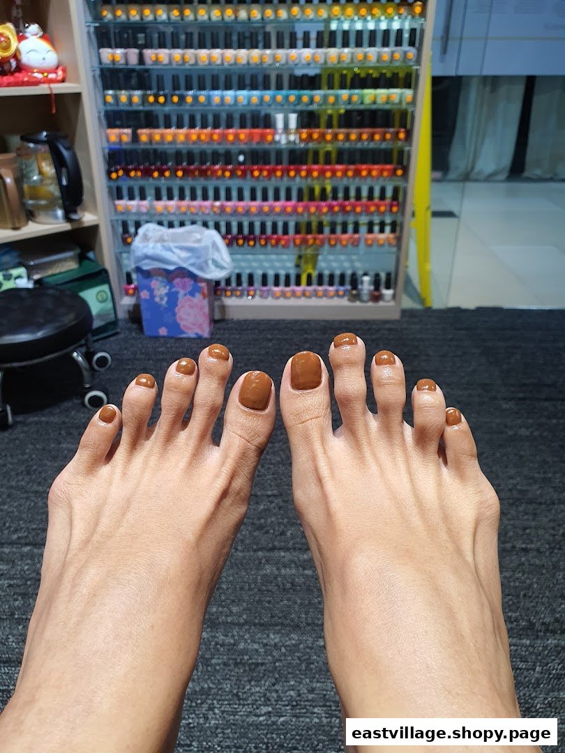 A close-up of feet with freshly painted brown toenails, with a large display of colorful nail polishes in the background.