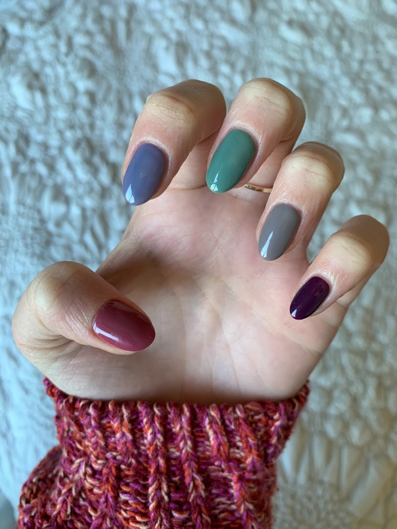 A hand showcasing five nails painted in different, complementary shades of blue, green, grey, and purple.