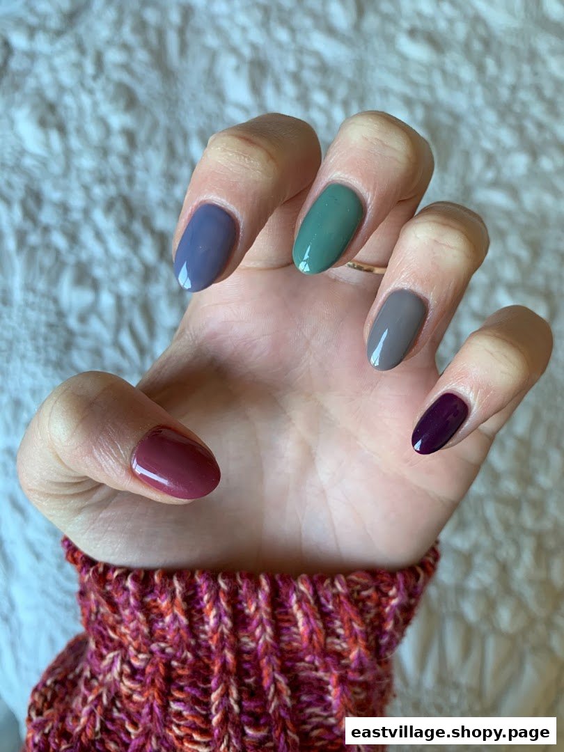 A hand showcasing five nails painted in different, complementary shades of blue, green, grey, and purple.