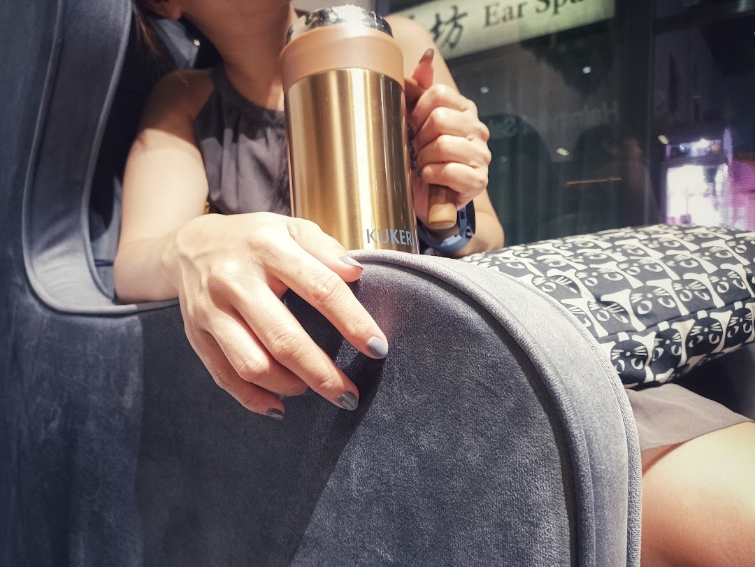 A person with manicured nails relaxes in a plush chair, holding a golden bottle, suggesting comfort and care.