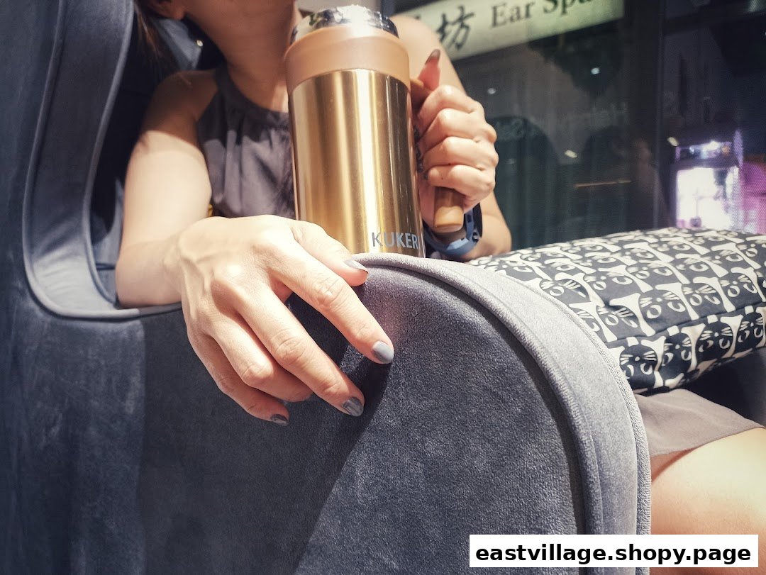 A person with manicured nails relaxes in a plush chair, holding a golden bottle, suggesting comfort and care.