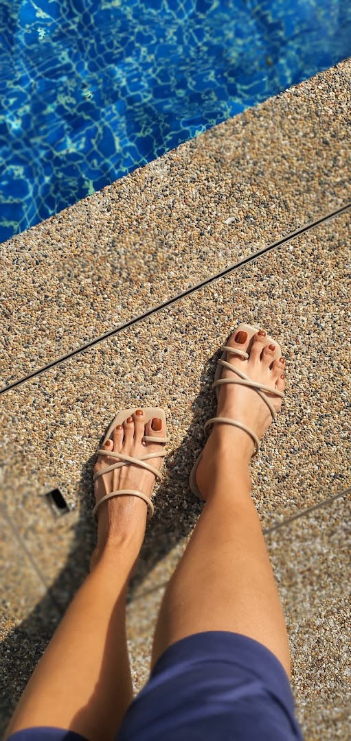 A person's feet with perfectly painted toenails and sandals by a sparkling blue swimming pool.