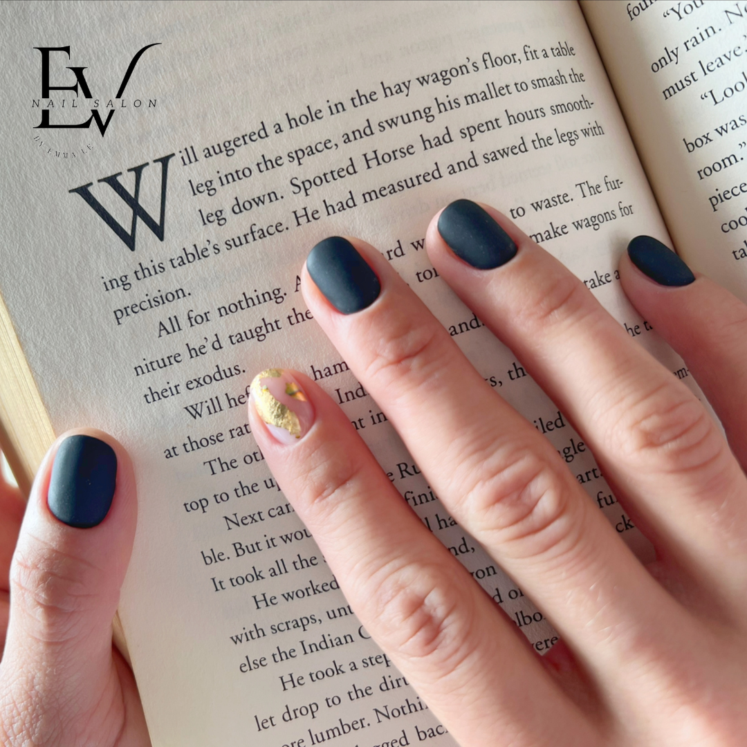 A hand with stylish dark matte nails and a gold foil accent nail holds an open book, featuring the salon's logo.