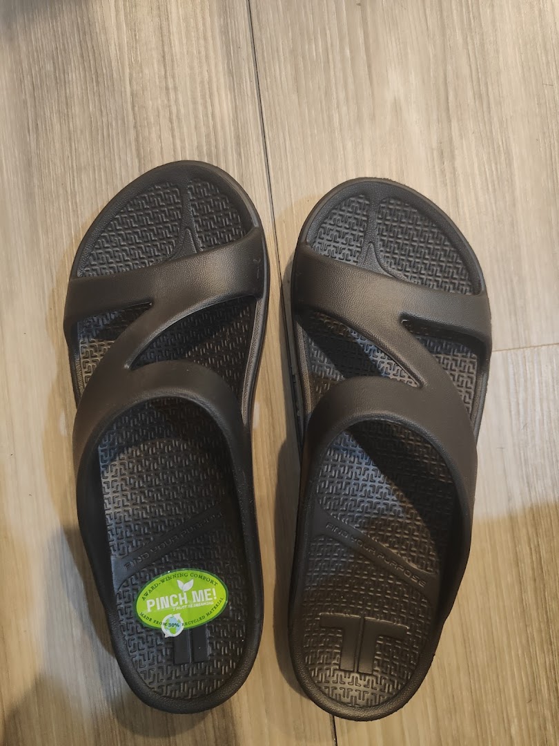 A pair of dark grey comfort sandals with a green 'Pinch Me!' sticker on a wooden floor.