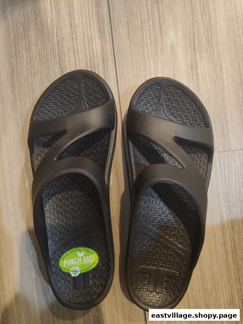 A pair of dark grey comfort sandals with a green 'Pinch Me!' sticker on a wooden floor.