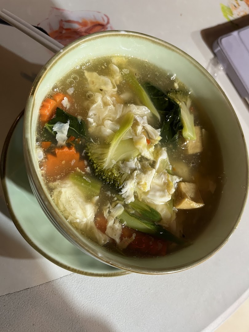 A close-up of a vibrant bowl of Thai soup with vegetables, tofu, and egg, served with chopsticks.
