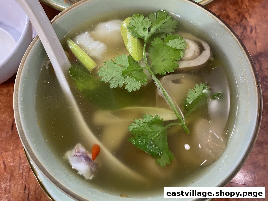 A close-up of a clear Thai soup with herbs, mushrooms, and meat in a light green bowl.