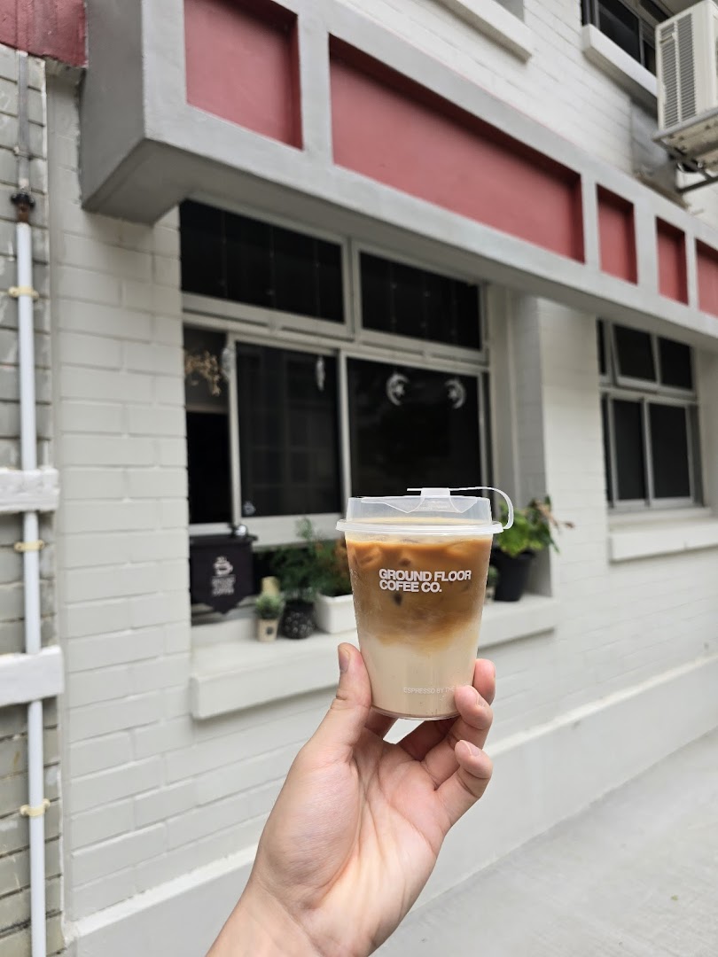 A hand holds an iced coffee cup from Ground Floor Coffee Co. in front of a white building.