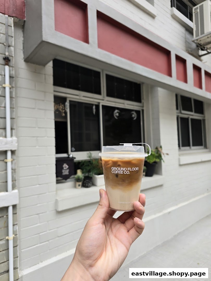 A hand holds an iced coffee cup from Ground Floor Coffee Co. in front of a white building.