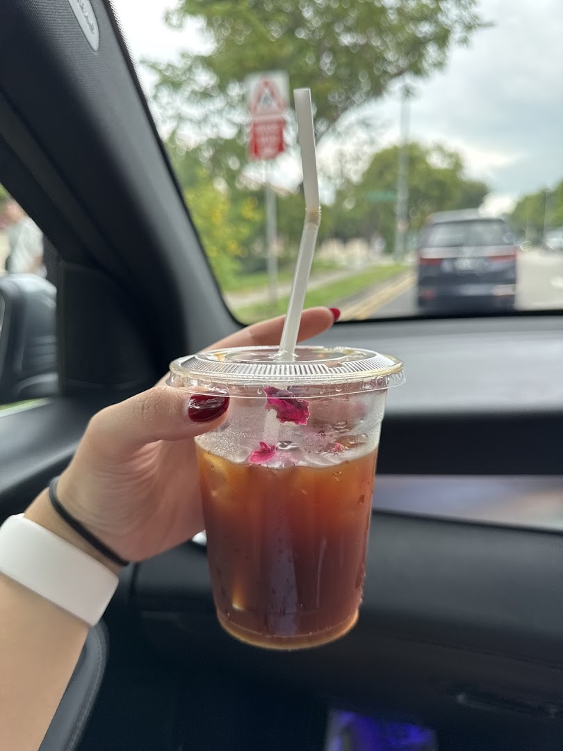 A hand holds an iced coffee with rose petals inside a car, with a street view in the background.