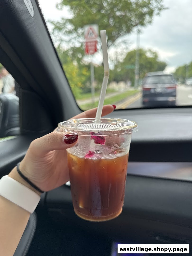 A hand holds an iced coffee with rose petals inside a car, with a street view in the background.