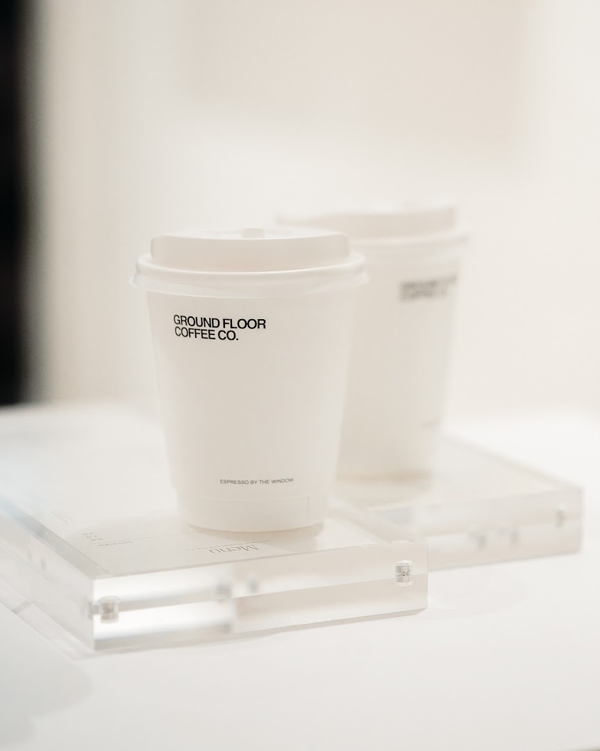 Two minimalist white coffee cups from Ground Floor Coffee Co. displayed on a clear stand.