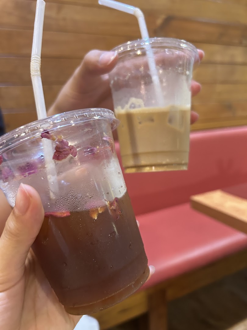 Two hands hold refreshing iced drinks, one coffee and one tea with rose petals, inside a cozy cafe.