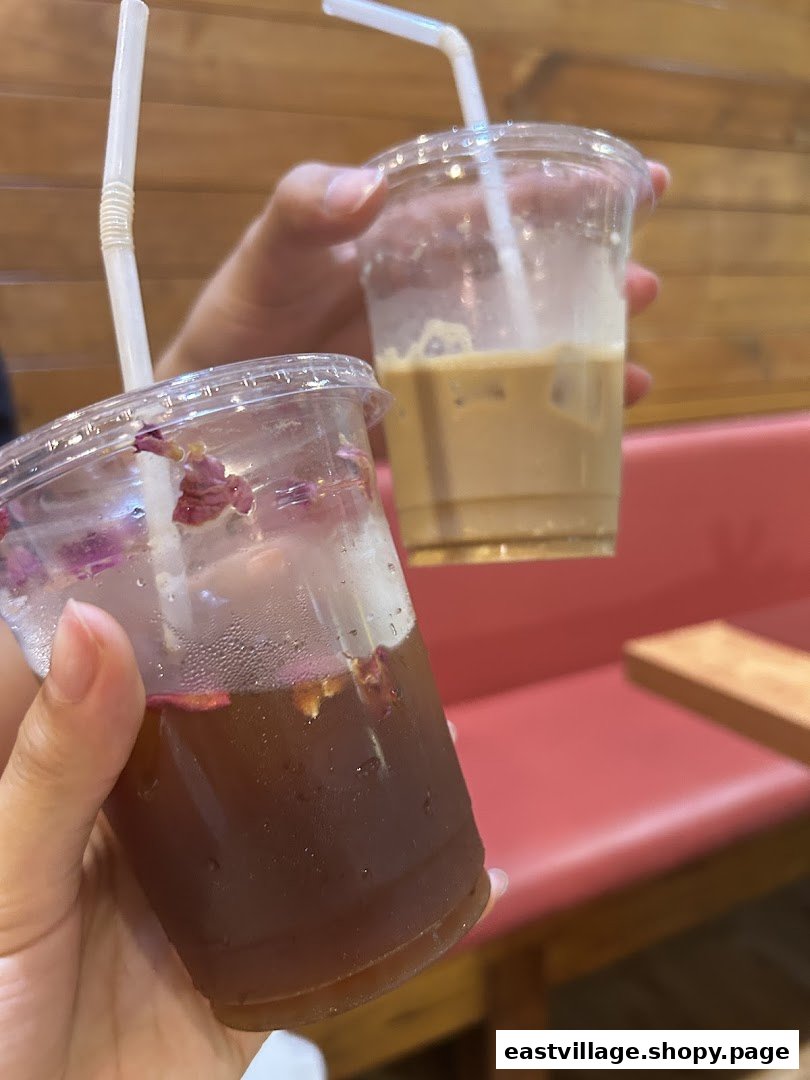 Two hands hold refreshing iced drinks, one coffee and one tea with rose petals, inside a cozy cafe.