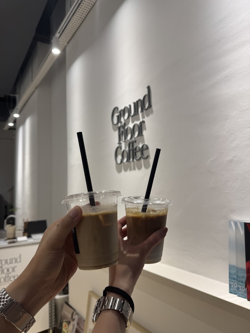Two hands hold iced coffee cups inside Ground Floor Coffee, with the shop's name on the wall.