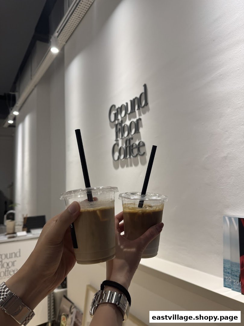 Two hands hold iced coffee cups inside Ground Floor Coffee, with the shop's name on the wall.