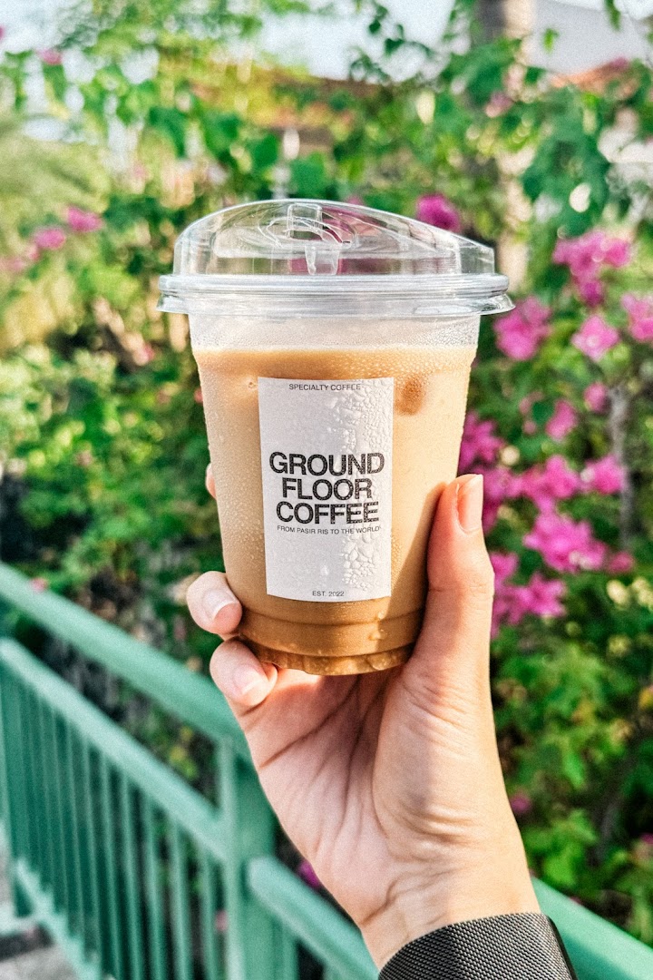 A hand holds an iced coffee cup from Ground Floor Coffee against a vibrant green and pink floral background.
