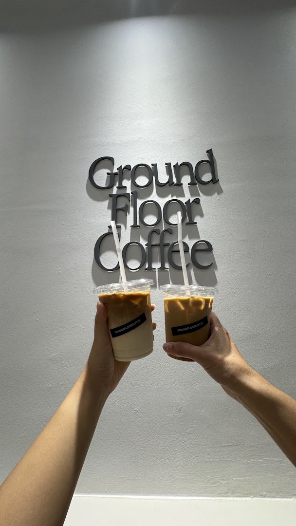 Two hands hold iced coffee cups in front of the 'Ground Floor Coffee' wall sign.