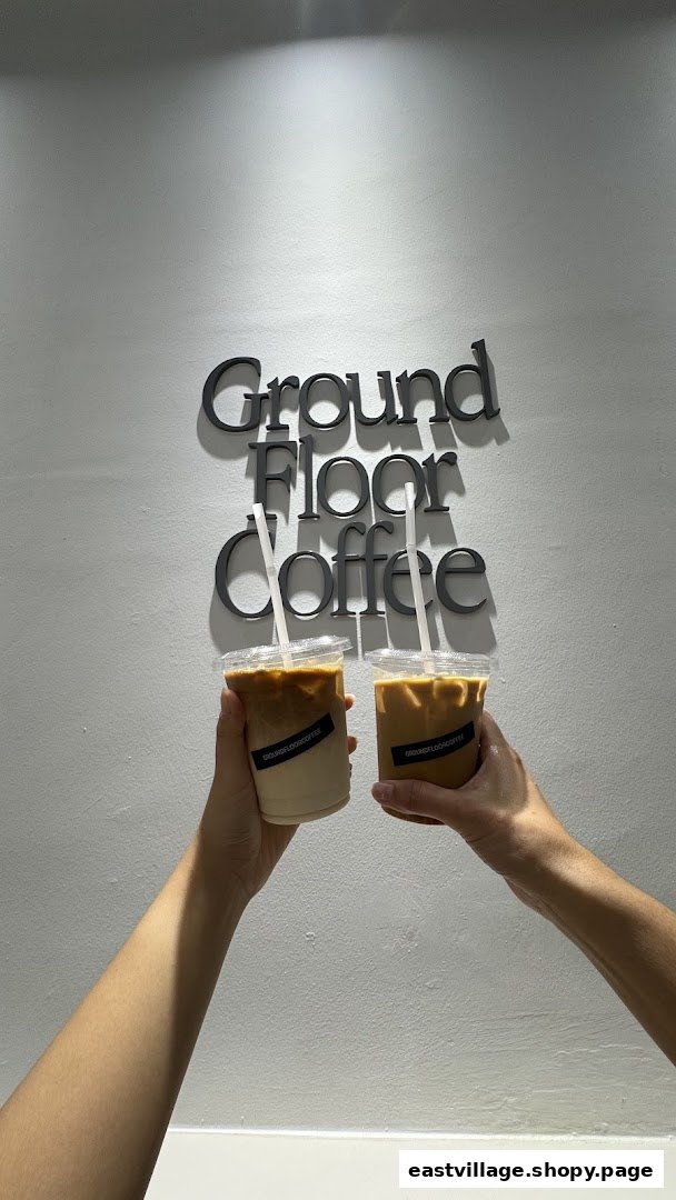 Two hands hold iced coffee cups in front of the 'Ground Floor Coffee' wall sign.