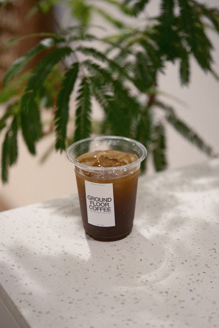 A refreshing iced coffee in a branded cup from Ground Floor Coffee, set on a light counter with green foliage.