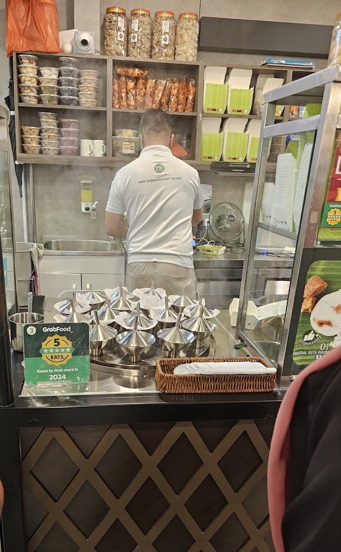 A staff member prepares food in a bustling kitchen, surrounded by packaged snacks and steaming putu piring molds.