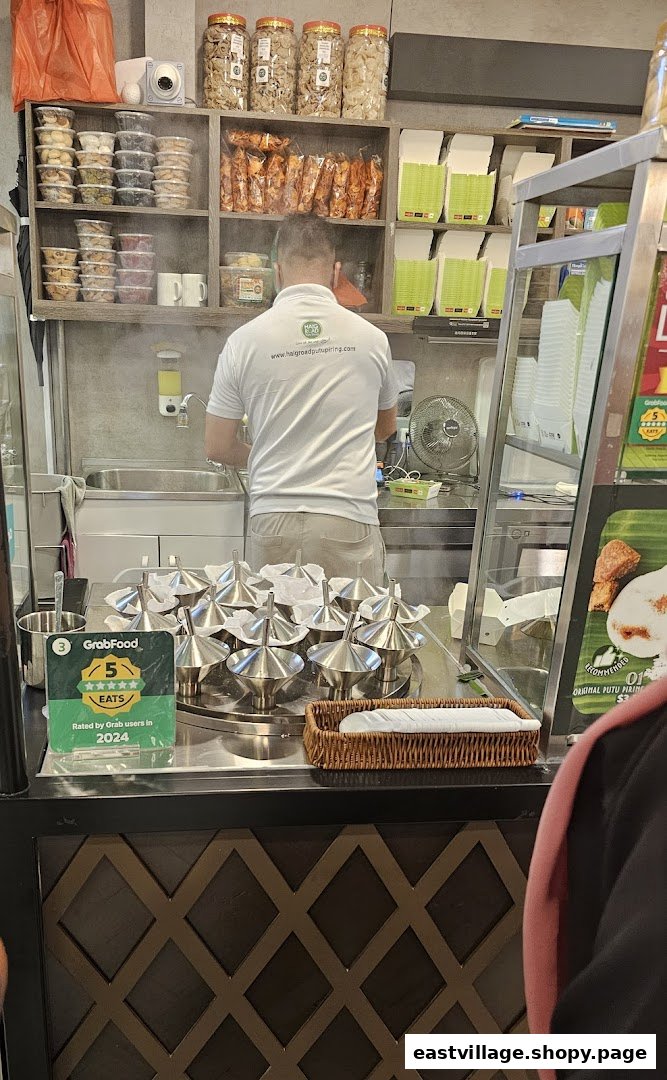 A staff member prepares food in a bustling kitchen, surrounded by packaged snacks and steaming putu piring molds.