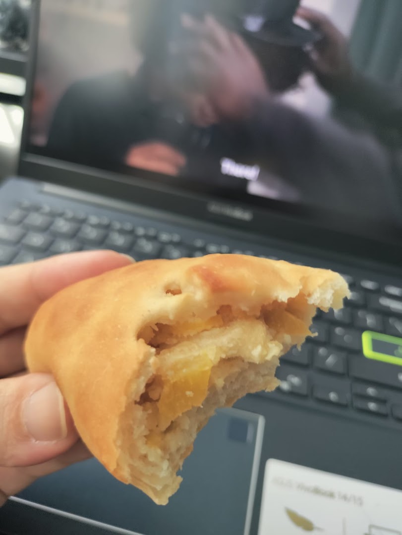 A hand holds a delicious, partially eaten bun with a sweet filling, in front of a laptop.
