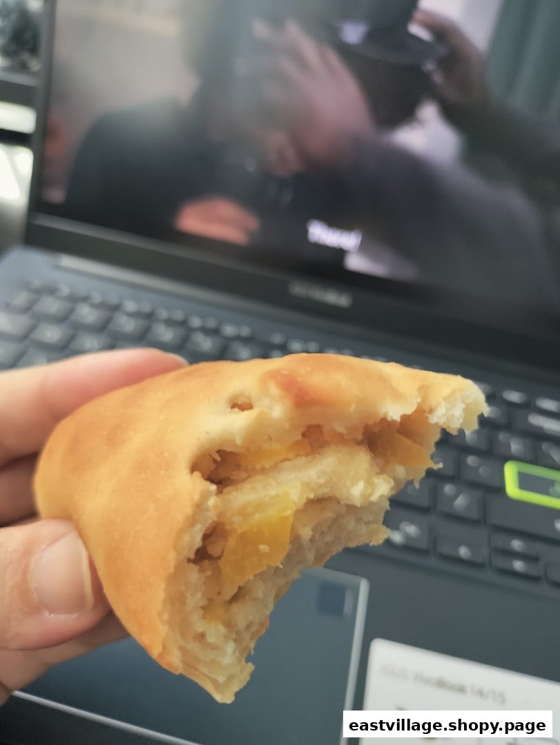 A hand holds a delicious, partially eaten bun with a sweet filling, in front of a laptop.