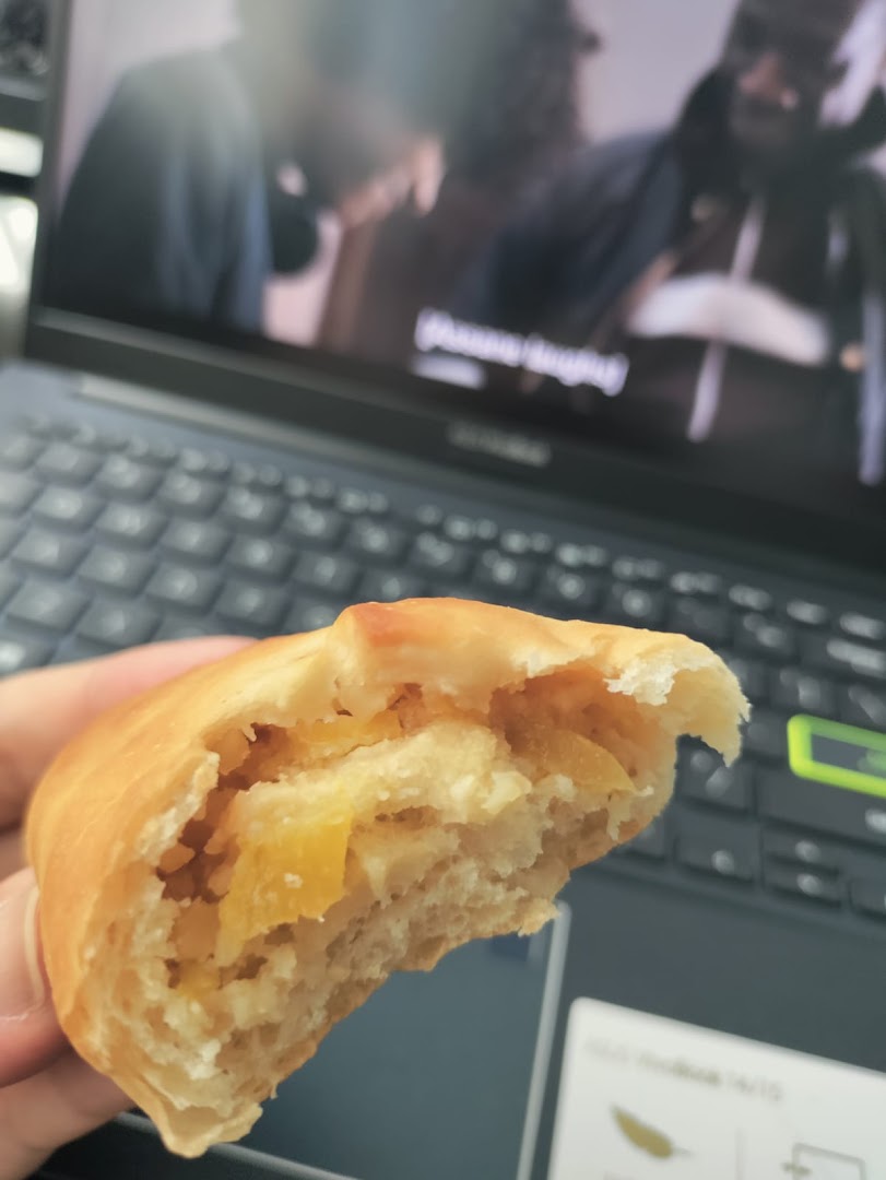 A hand holds a partially eaten, golden-baked bun revealing a sweet, yellow fruit filling.