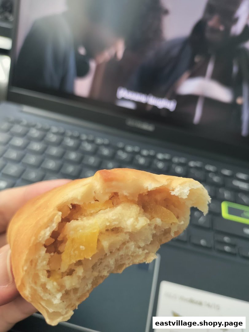 A hand holds a partially eaten, golden-baked bun revealing a sweet, yellow fruit filling.